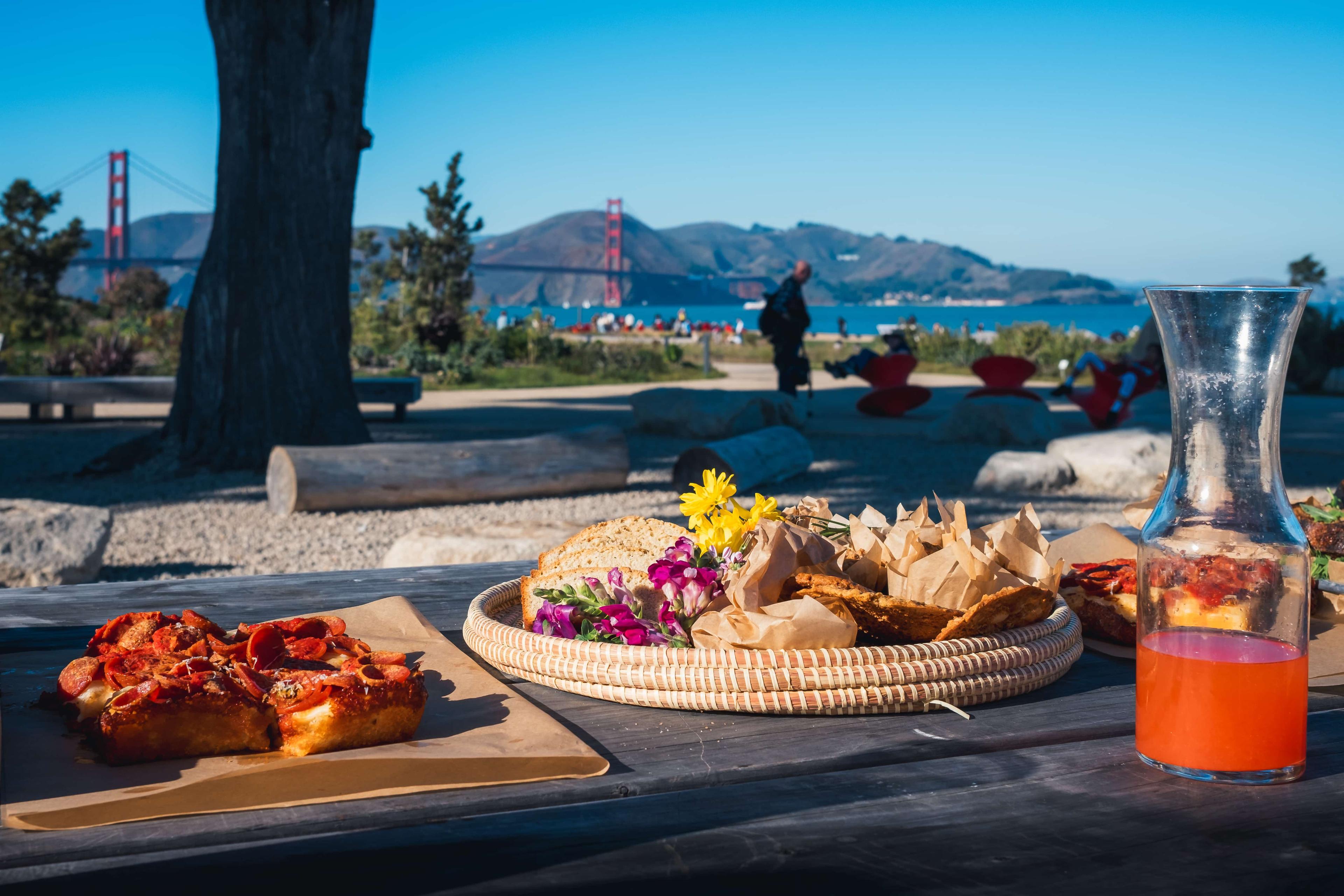 Picnic food platter with pizza and bread in the foreground and Presidio Tunnel Tops and the Golden Gate Bridge in the background.